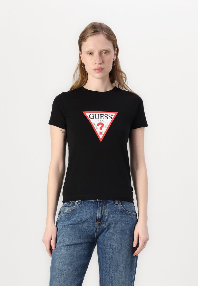 T-shirt en coton noir avec un logo triangulaire rouge audacieux portant le texte "GUESS ?". Manches courtes et coupe classique. Un jean en denim bleu complète le look.
