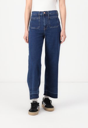 Person trägt hochgeschnittene, weite blaue Jeans mit großen Fronttaschen, weiße Socken, schwarze Sneaker und ein ärmelloses weißes Top.