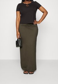 Vrouw in een zwart T-shirt met korte mouwen en kruisdetails, een olijfgroene lange rok, zwarte sandalen en een kleine zwarte handtas.