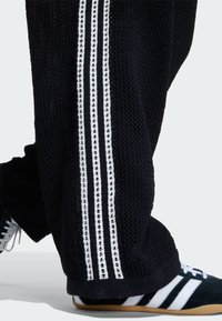Čierne pletené nohavice s bielymi bočnými pruhmi, ktoré obsahujú malé logá. Navrhnuté s voľným strihom a rebrovanými manžetami.