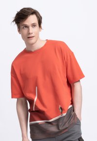 T-shirt en coton rouge à coupe décontractée. Présente un graphique d'une petite figure au niveau de l'ourlet, se fondant dans une section inférieure plus sombre et texturée.