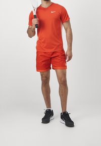 Červené sportovní tričko s výstřihem do V a bílým logem, spárované s červenými šortkami s vzorem. Černé sportovní boty s bílými akcenty, držící tenisovou raketu.