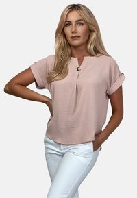 Jasnoróżowa bluzka z krótkimi, podwiniętymi rękawami i dekoltem w kształcie litery V, z dekoracyjnym złotym guzikiem na każdym rękawie. Noszona z białymi spodniami.