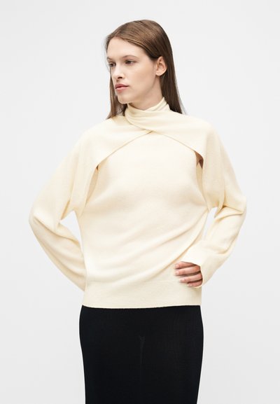 Γυναίκα με ίσια καστανά μαλλιά που φοράει εκρού πουλόβερ με ψηλό λαιμό και προσαρμοσμένο ντραπαριστό σάλι, καθώς και μαύρη φούστα, με τα χέρια στους γοφούς.