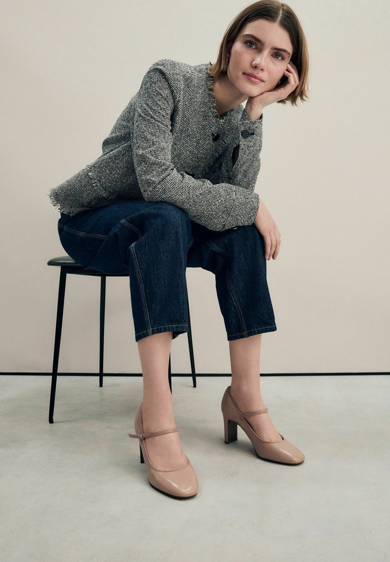 Zapatos de tacón alto de charol beige con una correa en la parte frontal, combinados con una chaqueta gris texturizada y unos pantalones vaqueros oscuros cropped.