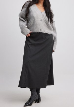 Femme portant un cardigan gris boutonné, une jupe mi-longue gris foncé, des chaussettes grises et des mocassins noirs à talons, se tenant devant un fond uni.