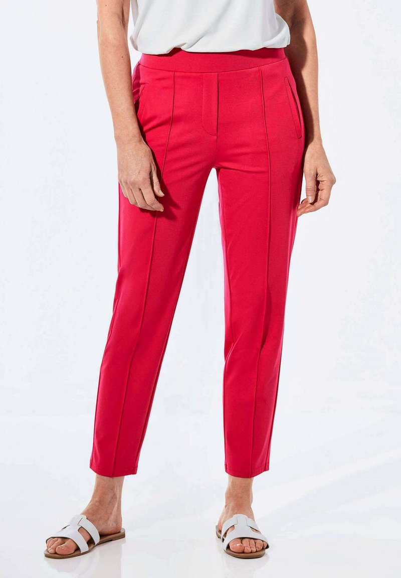 Rote maßgeschneiderte Hosen mit glatter Textur, ausgestattet mit vorderen Taschen und dezenten Falten. Getragen mit einem weißen Oberteil und hellen Sandalen.