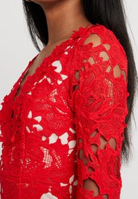 Robe en dentelle rouge avec des motifs floraux, un décolleté en V profond et des détails découpés élaborés sur les manches, mettant en valeur la texture et le design.