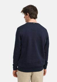 Mørkeblå genser med rund hals, ribbestrikkede ermer og kant. Har en liten logo-tag på baksiden av halsen. Myk tekstur.