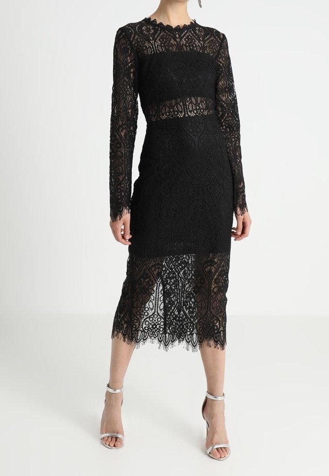Femme portant une robe midi noire en dentelle à manches longues avec des panneaux transparents et des sandales à talons hauts argentées, debout devant un fond uni.