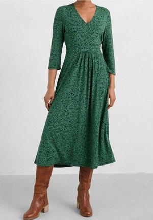 Femme portant une robe midi verte à motifs avec des manches trois-quarts et des bottes brunes à talons jusqu'aux genoux, debout sur un sol gris.