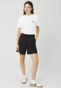 T-shirt graphique blanc avec le texte "Sweet or Sour". Shorts noirs cintrés avec plis et poches latérales. Le mannequin porte des baskets blanches avec des accents noirs.