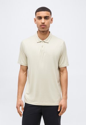 Noor mees lühikeste juustega, kellel on seljas beež lühikeste varrukatega polo-särk ja mustad püksid, seisab helehalli tausta ees.