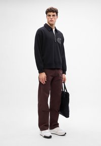 Felpa nera con zip e colletto, con logo sul petto; abbinata a pantaloni bordeaux e scarpe da ginnastica bianche. Tiene in mano una borsa nera.