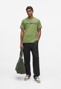 Camiseta de algodón verde con logo negro "KARL LAGERFELD", combinada con pantalones negros, zapatos beige y oscuros, y un bolso tote verde.