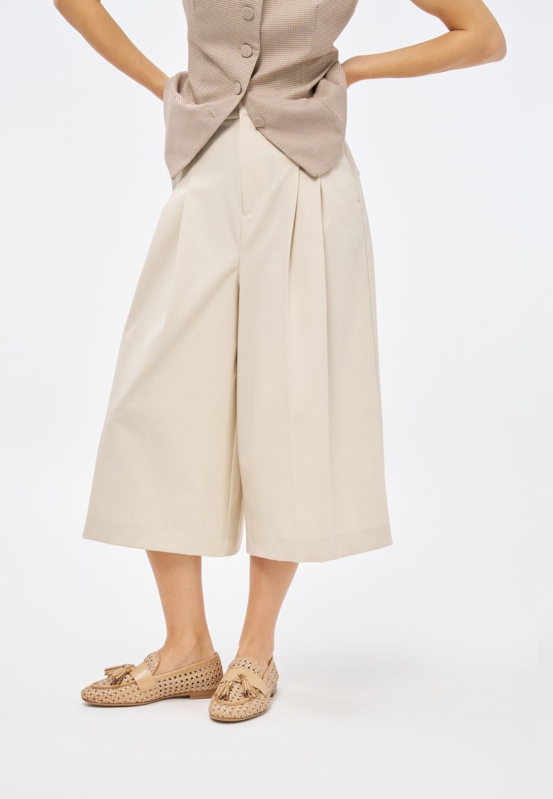 Pantaloni beige a gamba larga con pieghe, realizzati in tessuto leggero. Indossati con scarpe slip-on beige in tessuto intrecciato con dettagli a frange.