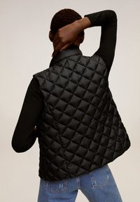 Černá prošívaná puffer vesta s vysokým límcem. Texturovaný diamantový vzor, lehký materiál. Nositá přes dlouhý rukáv a džíny.