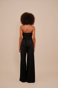 Kobieta z kręconymi włosami, nosząca bezrękawowy czarny kombinezon z kokardą z tyłu, stojąca na tle jednolitej beżowej ściany, odwrócona plecami.