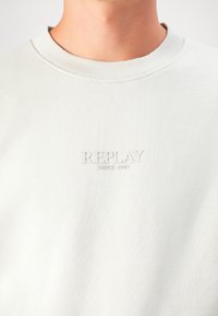 Close-up of a person wearing a white sweatshirt with a ribbed crew neck and raised "REPLAY SINCE 1981" text on the chest.