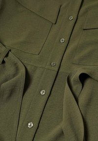 Gros plan sur un tissu vert olive avec une fermeture à boutons sur le devant et deux poches poitrine rectangulaires avec rabats.