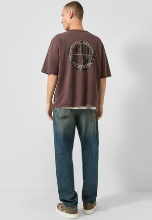 Man draagt een bruine oversized T-shirt met een cirkelvormige afbeelding en tekst op de achterkant, blauwe jeans en bruine sneakers, staand tegen een effen achtergrond.