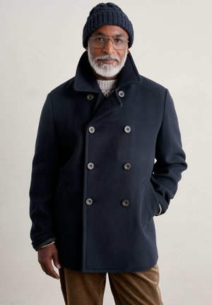 Cappotto peacoat di lana navy con bottoni doppi, grande colletto e tasche laterali, abbinato a un berretto di maglia scura a coste e pantaloni marroni strutturati.