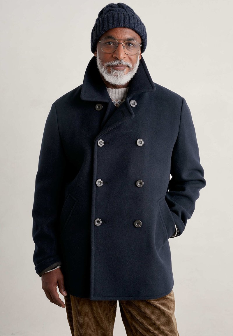 Cappotto peacoat di lana navy con bottoni doppi, grande colletto e tasche laterali, abbinato a un berretto di maglia scura a coste e pantaloni marroni strutturati.