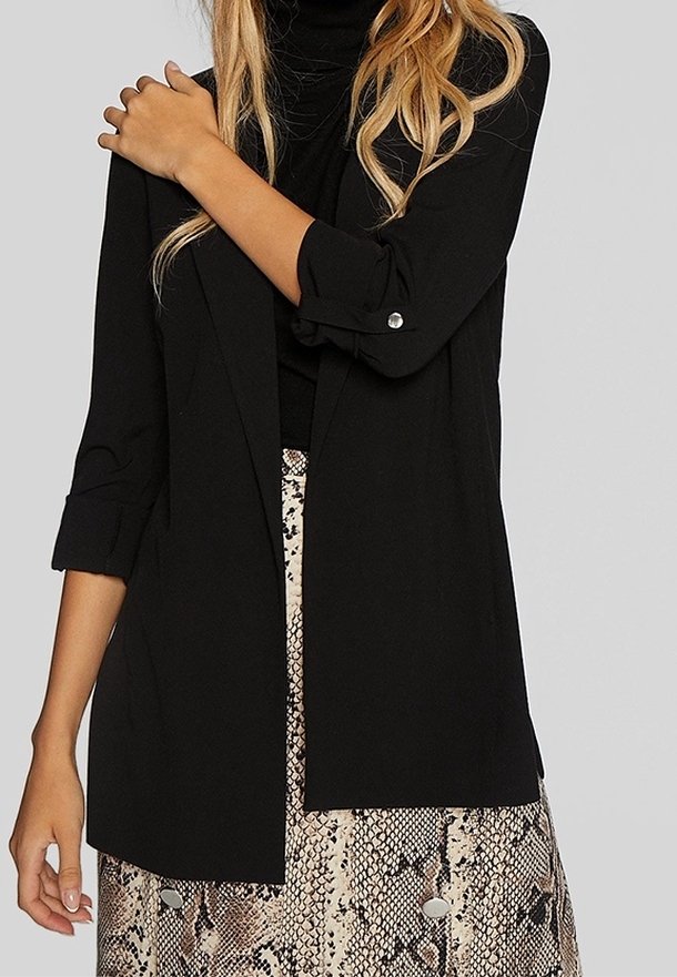 Femme portant un blazer noir à manches longues sur un top noir, associé à une jupe beige et marron imprimé serpent.