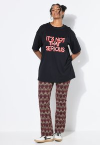 Zwarte oversized t-shirt met rode grafische tekst "HET IS NIET ZO ERG," gecombineerd met aansluitende geblokte broek in bordeauxrood en beige, afgewerkt met sneakers.