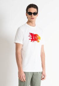 Camiseta de algodón blanca con un gráfico de león en rojo y naranja. El modelo lleva gafas de sol negras y pantalones verdes claros.