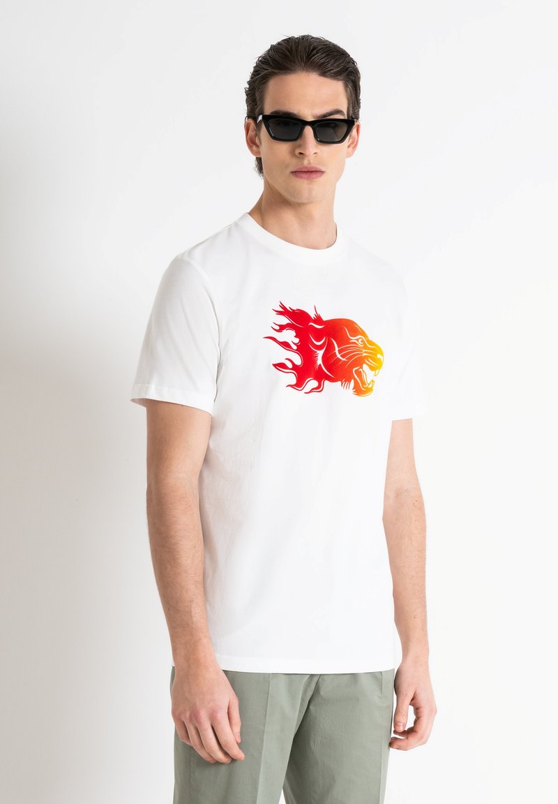 Camiseta de algodón blanca con un gráfico de león en rojo y naranja. El modelo lleva gafas de sol negras y pantalones verdes claros.