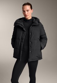 Fekete puffajak, kapucnival, gombos elülső résszel, steppelt textúrával és oldalsó zsebekkel, fekete, szűk leggingsszel párosítva.