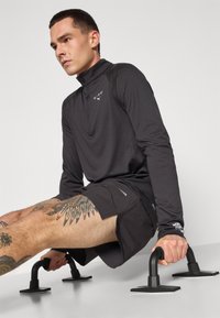 Svart langermet treningsgenser med glidelås og reflekterende logo, kombinert med svarte shorts. Inkluderer ergonomisk utformede push-up-stenger.