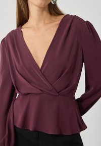 Blusa envolvente burdeos con escote en V profundo, hombros plisados y dobladillo en peplum, confeccionada en una tela suave y ligera.