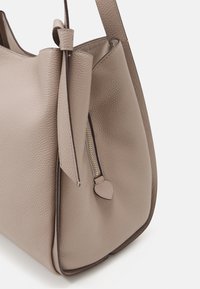 Beige kožená kabelka s texturovaným povrchem, obsahující zlatý zip v kapse a uvázaný detail na popruhu. Zaoblený tvar s minimálními akcenty.