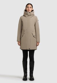 Beige buitenjack met een hoge kraag en capuchon, voorzien van twee voorzakken, een ritsluiting en een aansluitende pasvorm. Gedragen boven zwarte leggings en kniehoge laarzen.