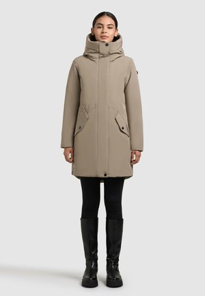 Giacca esterna beige con colletto alto e cappuccio, dotata di due tasche frontali, chiusura con zip e una vestibilità sfiancata. Indossata sopra leggings neri e stivali fino al ginocchio.