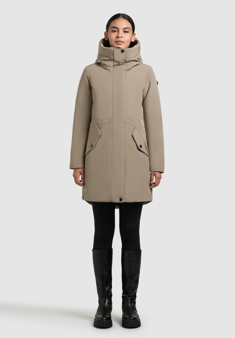 Beige buitenjack met een hoge kraag en capuchon, voorzien van twee voorzakken, een ritsluiting en een aansluitende pasvorm. Gedragen boven zwarte leggings en kniehoge laarzen.