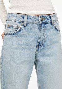 Ljusblå denimjeans har hög midja, femficksdesign och subtila urtvättning. Grått stitching och metallknappdetaljer är synliga.