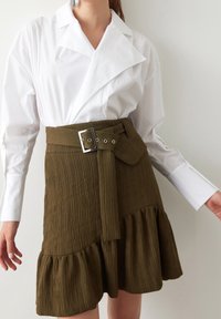 Camisa blanca con cuello, superpuesta a una falda texturizada verde caqui. La falda cuenta con una cintura con cinturón y un dobladillo con volantes.