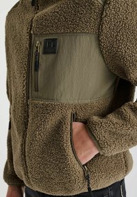 Bruine fleece jas met een getextureerde buitenkant, olijfgroene paneel, borstzak en verstelbare trekkoord aan de zoom. Gehoogd logo detail zichtbaar.