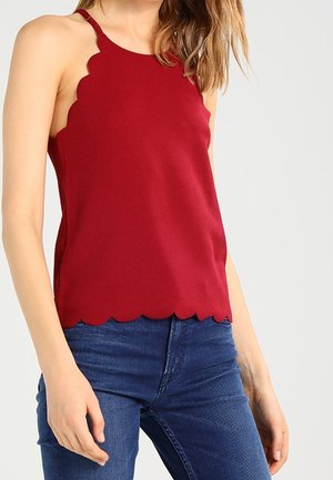 Kvinde iført en ærmeløs rød top med bølget kant og blå jeans mod en ensfarvet lys baggrund.