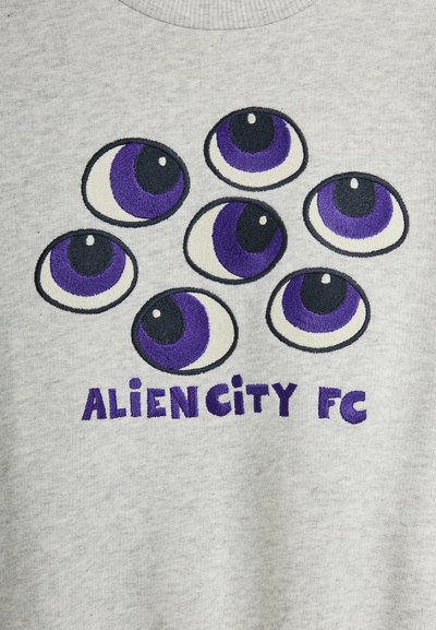 Επτά μωβ και μαύρα κεντημένα μάτια διατεταγμένα πάνω από το κείμενο «ALIEN CITY FC» σε ανοιχτό γκρι ύφασμα.