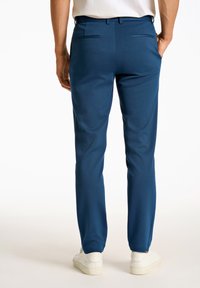 Blaue, maßgeschneiderte Hosen mit glatter Textur, zwei Gesäßtaschen und schlanker Passform, kombiniert mit weißen Sneakers vor einem hellen Hintergrund.