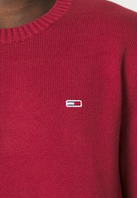 Maglione rosso lavorato a maglia con scollatura a coste. Presenta un piccolo logo bianco, blu e rosso sul petto, che mette in evidenza una texture liscia.