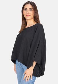 Schwarzer, leichter Poncho mit kastenförmiger Silhouette, strukturiertem Stoff und Spitze am Saum, kombiniert mit blauen Jeans.