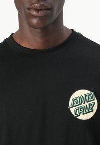 Black cotton t-shirt with a round logo patch featuring the text "SANTA CRUZ" in green on the left chest area. Short sleeves, classic neckline.
