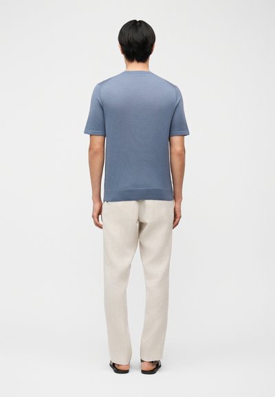 Άνδρας όρθιος με την πλάτη, φοράει κοντομάνικο μπλε πουκάμισο, ανοιχτό μπεζ παντελόνι και μαύρα σανδάλια σε απλό λευκό φόντο.