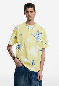 Sárga pamut póló kék virágmintákkal, kerek nyakkivágással, rövid ujjakkal és laza szabással. Látható logók nélkül, csak egy apró kék részlettel.