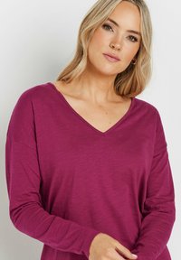 Langärmliges V-Ausschnitt-Top in tiefem Magenta, aus weichem Stoff mit lockerer Passform und subtiler Textur. Mit herunterhängenden Schultern.